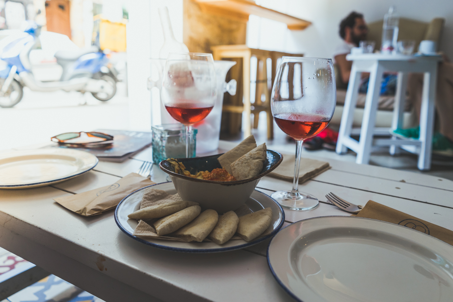 Een witte tafel gedekt voor twee met glazen rosé, borden, servetten en broodstengels wijst op een gezellige srprs.me citytrip. Zonlicht valt naar binnen terwijl een blauwe scooter buiten wacht en iemand aan een nabijgelegen tafel van zijn dag geniet.