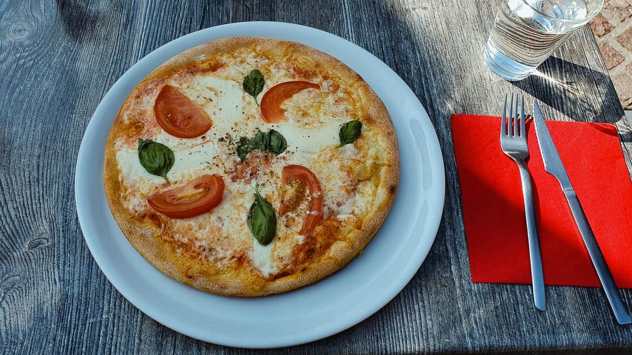 Een Margherita pizza met plakjes tomaat, mozzarella kaas en basilicumblaadjes ligt op een wit bord op een rustieke houten tafel. Ernaast ligt keukengerei op een rood servet en een glas water-perfecte gastvrije verwennerij na een citytrip Ronse.