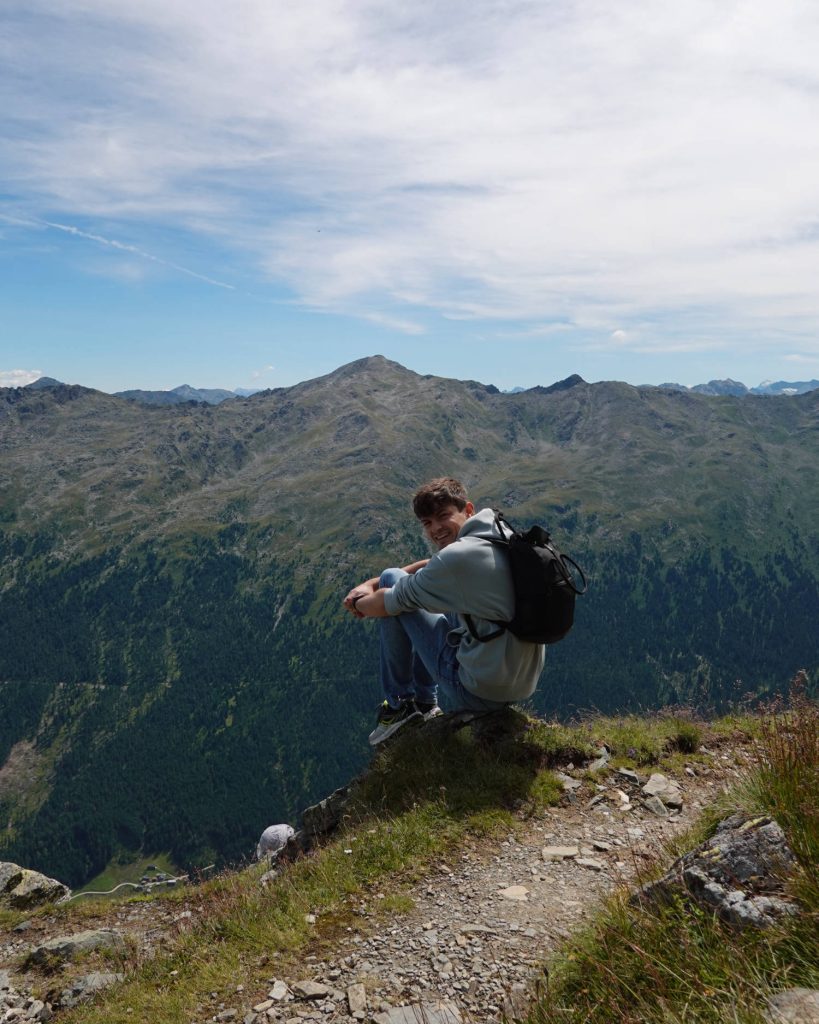 Een persoon met een rugzak zit aan de rand van een bergpad, glimlacht naar de camera, met een schilderachtig uitzicht op groene valleien en ruige bergtoppen onder een gedeeltelijk bewolkte hemel op de achtergrond.