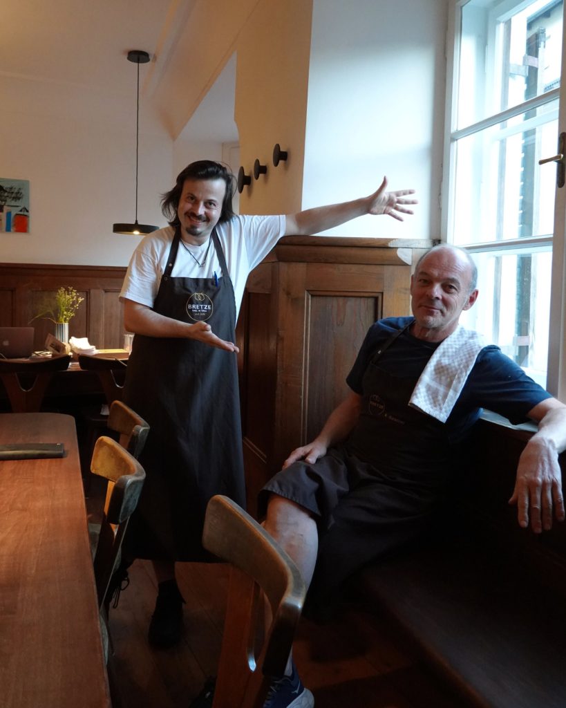 Twee mannen in schorten zitten in een gezellig restaurant in Tirol. De een staat glimlachend en gebarend naar de ander, die op een bankje bij een helder raam zit met een handdoek over zijn schouder. Houten tafels en stoelen vullen de ruimte.
