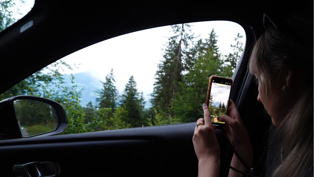 Iemand die in een auto in Tirol zit, maakt een foto van een schilderachtig, boomrijk landschap door het open raam, waarbij hij hoge evergreens en bergen in de verte vastlegt op zijn smartphone - perfect voor de zomerchecklist van dingen die hij nog moet doen.