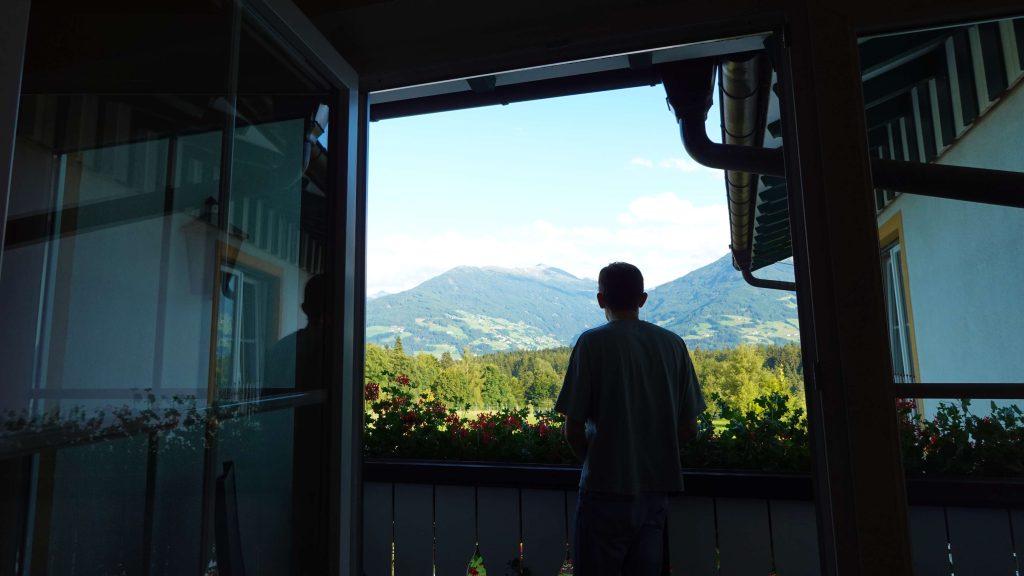 Iemand staat op een balkon in Tirol en kijkt uit over groene heuvels en bergen in de verte onder een helderblauwe hemel - misschien plant hij zijn zomerchecklist, omringd door een open deur en huismuren.