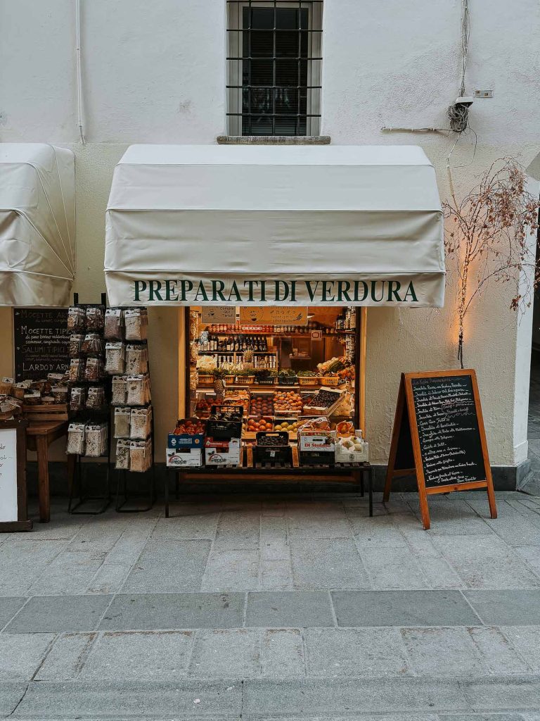 Een kleine groentewinkel met een witte luifel met de tekst "PREPARATI DI VERDURA," toont verse producten en verpakte goederen uit Valle d'Aosta. Rechts staat een krijtbordmenu en links hangen Bergenmagie-verpakkingen.