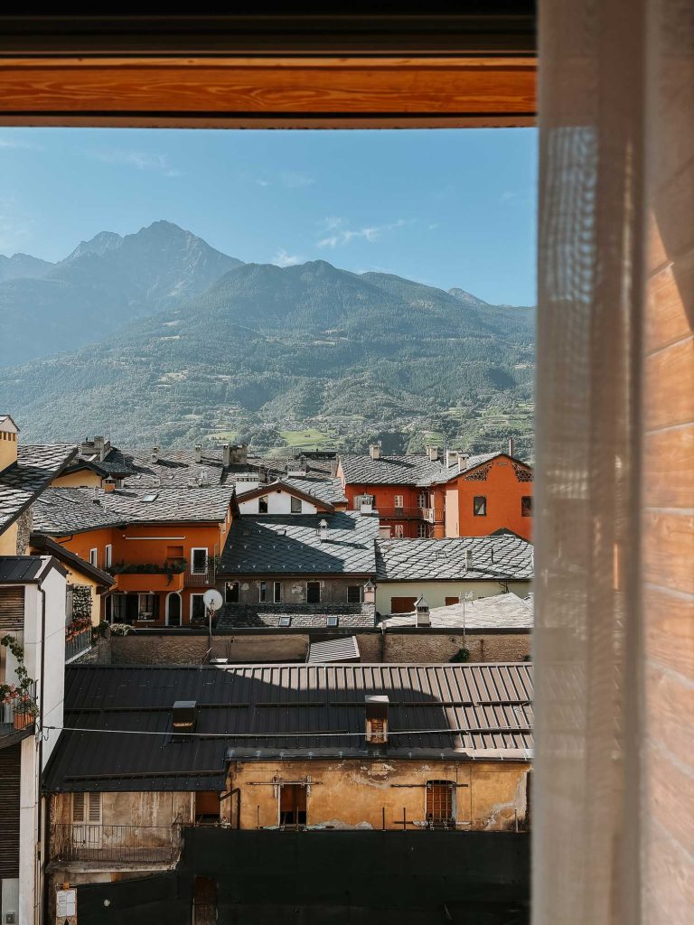Uitzicht vanuit een raam op kleurrijke daken van een dorp in Valle d'Aosta met rode en gele gebouwen tegen groene bergen en een helderblauwe lucht. Een gordijn is gedeeltelijk zichtbaar aan de rechterkant, wat bijdraagt aan de Bergenmagie charme.