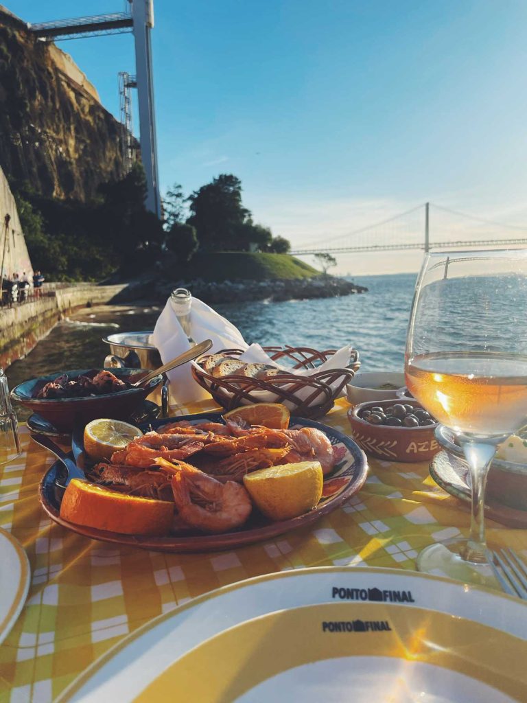 Een gedekte tafel aan zee met zeevruchten, brood en een glas roséwijn bij zonsondergang, met uitzicht op de rivier en de hangbrug van Lissabon. Dineren bij "Ponto Final" is een van de must-dos voor een onvergetelijke stadservaring.