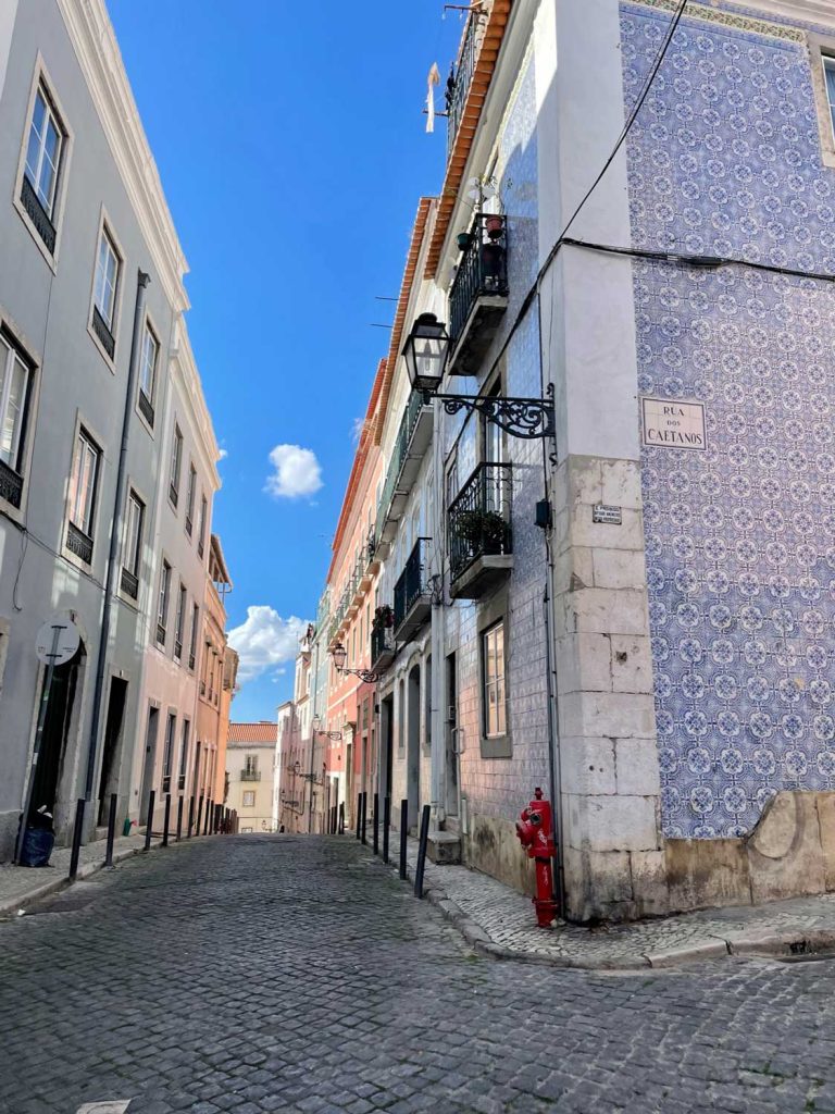 Een smalle geplaveide straat in Lissabon met oude gebouwen, waaronder een met blauwe tegels - een must voor bezoekers. Op een straatnaambordje staat "Rua Caetano" en op de hoek staat een rode brandkraan onder een strakblauwe hemel.