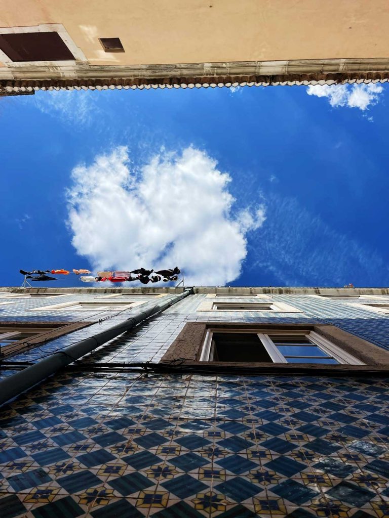 Als je omhoog kijkt tussen twee gebouwen met gedessineerde tegels in Lissabon, zie je daarboven een helderblauwe lucht en pluizige witte wolken. Kleren hangen aan een lijn voor een raam en voegen kleur toe aan deze lijst van must-dos voor stadsverkenners.
