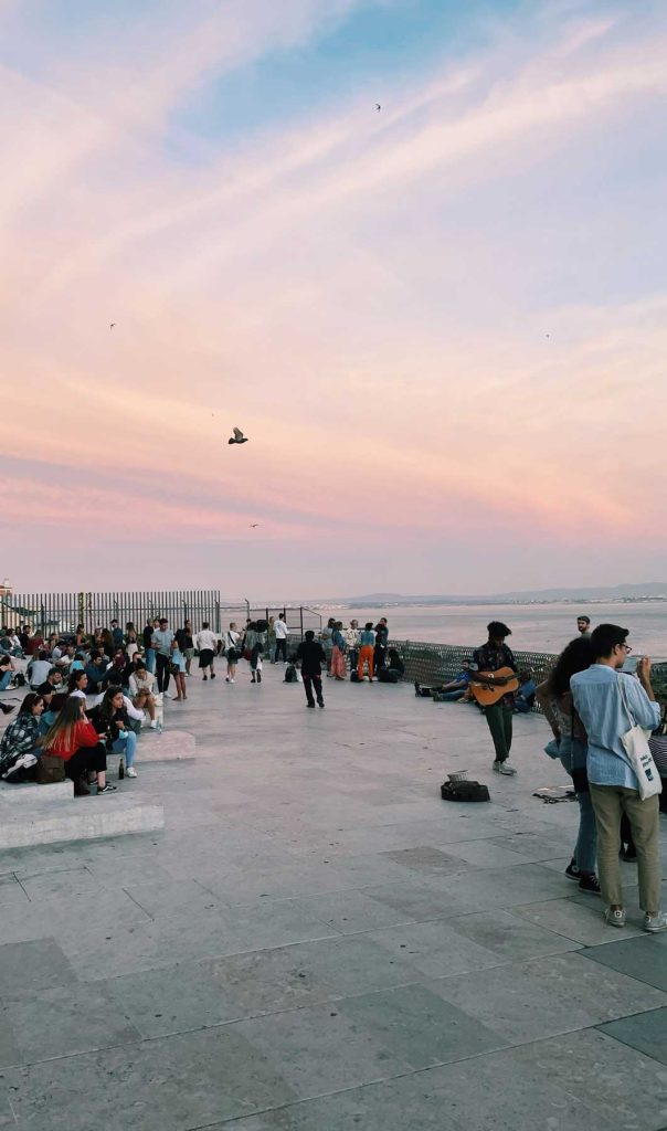 Mensen verzamelen zich op een promenade aan zee in Lissabon bij zonsondergang, sommigen zitten op bankjes en anderen staan. Een straatmuzikant speelt gitaar terwijl vogels in de pastelkleurige lucht vliegen - een levendig, ontspannen tafereel tussen de must-dos van de stad.