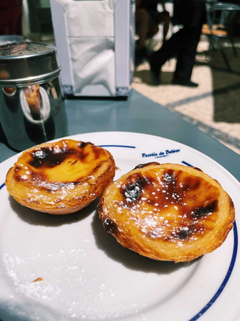 Twee Portugese custardtaartjes, bekend als pastéis de nata - een onmisbare traktatie in Lissabon - liggen op een wit bord met een blauwe rand, licht bestrooid met poedersuiker. Een metalen suikerstrooier en servethouder staan op de achtergrond.