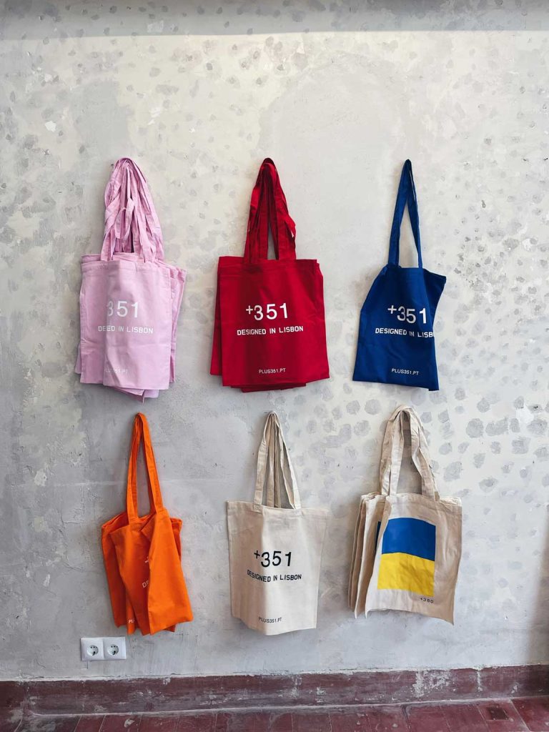 Zes draagtassen hangen aan een gestructureerde, lichtgrijze muur - perfect om je Lissabon must-dos in mee te nemen. De tassen zijn roze, rood, blauw, oranje, beige met tekst en beige met een blauw en geel ontwerp dat lijkt op de Oekraïense vlag. Onderaan is een stopcontact zichtbaar.