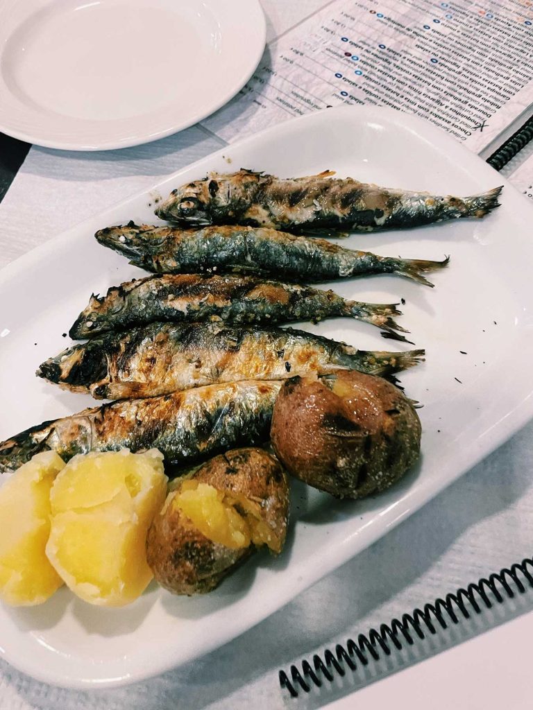 Vijf gegrilde sardientjes geserveerd op een wit bord met twee gekookte aardappelen en twee gepofte aardappelen - een klassieker in Lissabon - op een tafel met een wit tafelkleed en een menukaart op de achtergrond.