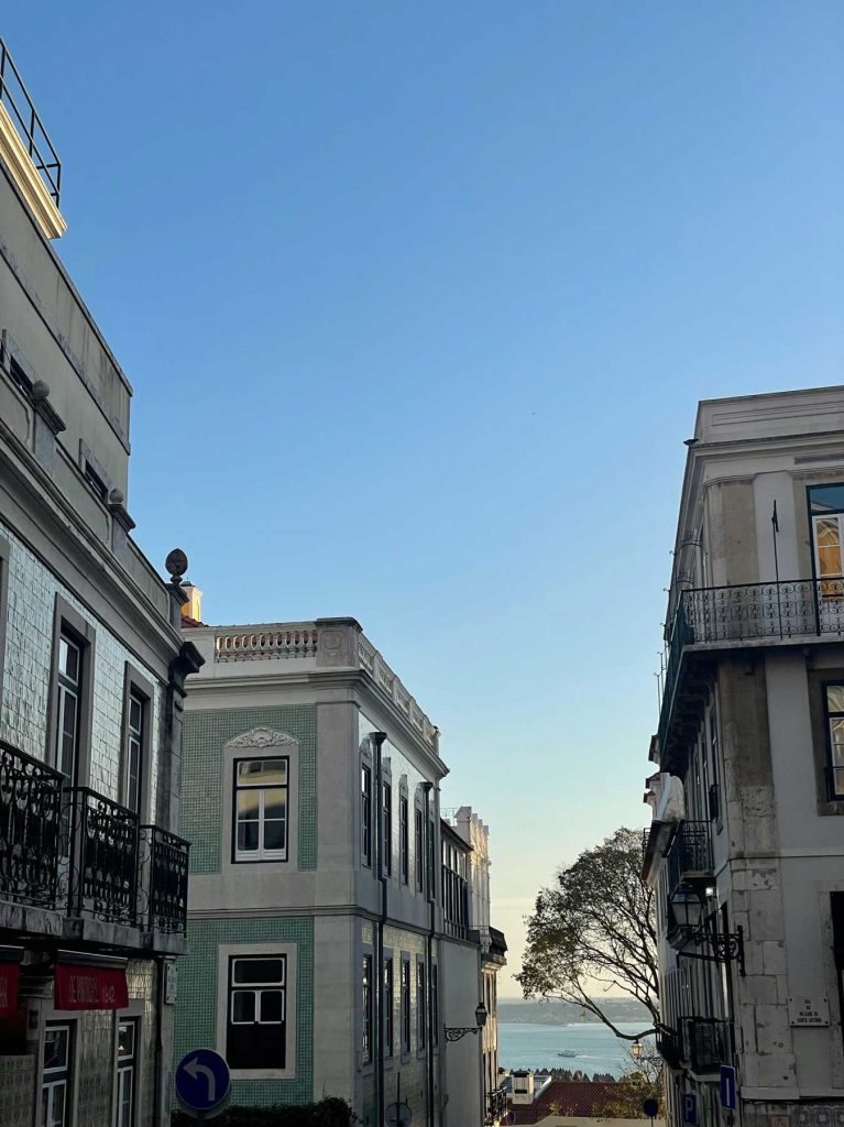 Een strakblauwe lucht boven een smalle straat met oude gebouwen en smeedijzeren balkons. In de verte zie je een glimp van water en een boom. Dit is de klassieke sfeer van Lissabon en een van de must-dos voor bezoekers.