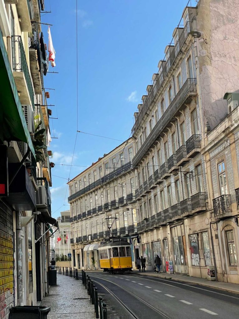 Een gele tram rijdt door een smalle straat met oude, sierlijke gebouwen in Lissabon, Portugal - een must voor bezoekers terwijl voetgangers over het trottoir wandelen onder een strakblauwe hemel.