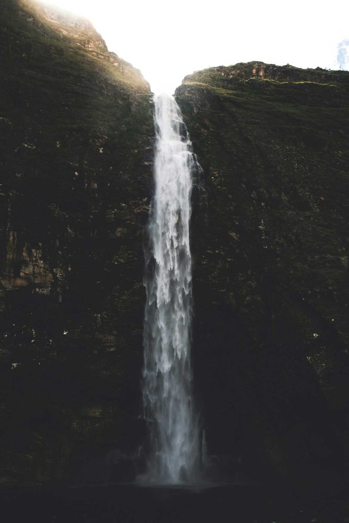 Een hoge, smalle waterval stort van een steile, rotsachtige klif omgeven door donkergroene vegetatie in de koffielanden, met zonlicht dat schijnt op de top waar het water begint te vallen.