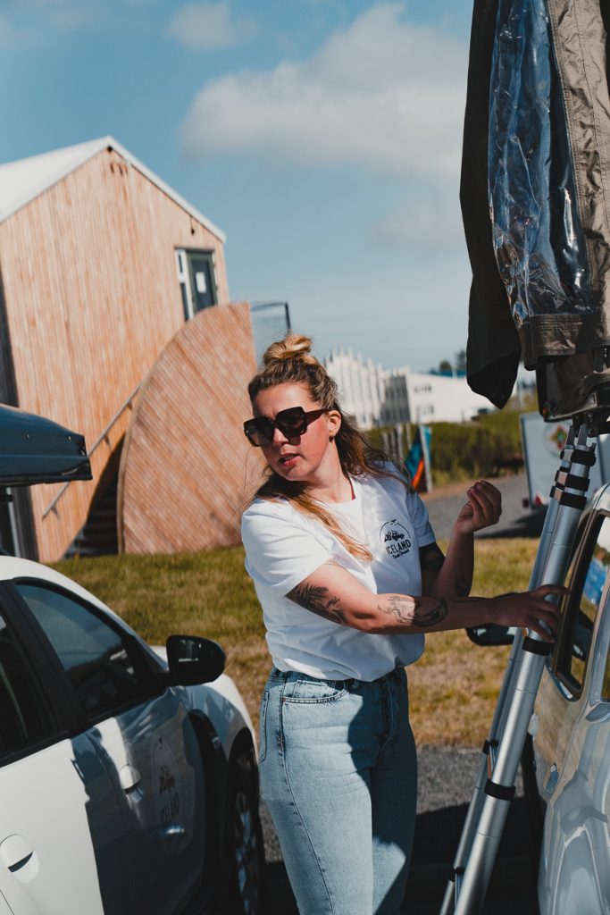 Een vrouw met een zonnebril, een wit T-shirt en een spijkerbroek staat naast een auto en past iets aan op een daktent - een perfect tafereel voor IJslandse nomaden. Houten gebouwen staan op de achtergrond en het weer lijkt zonnig.