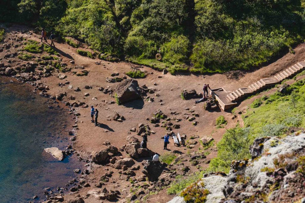 Luchtfoto van mensen die lopen en staan op een rotsachtig, aards gebied bij water in IJsland, met trappen die naar beneden leiden vanaf een groene heuvel - een inspirerende scène van natuurlijke groei en ontdekking.