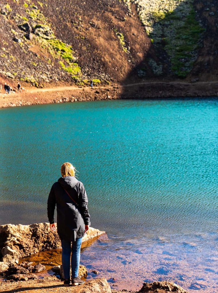 Een persoon in een donkere jas staat op rotsen aan de rand van een levendig blauw meer in IJsland, omgeven door steil, rotsachtig terrein met plekken mos en zonlicht die de serene groei van de natuur benadrukken.