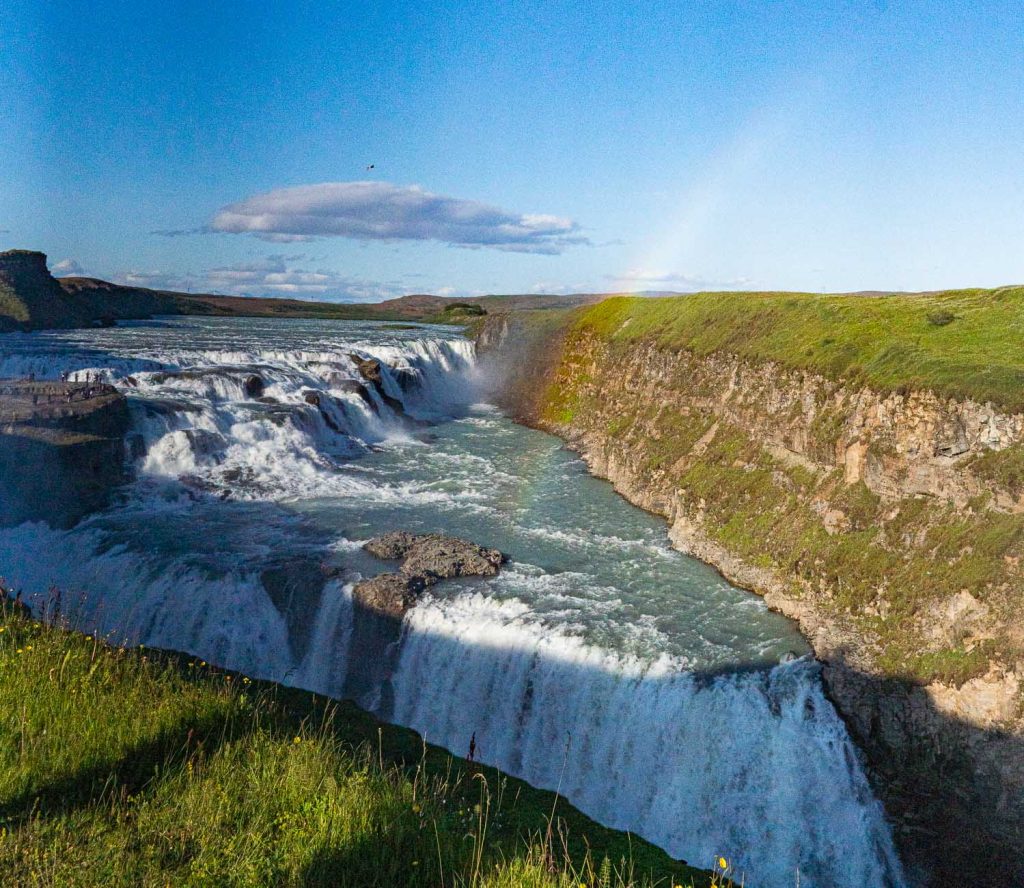 Een krachtige waterval in IJsland stort zich over rotsachtige kliffen in een rivier, met een regenboog vaag zichtbaar in de mist onder een blauwe hemel. Weelderig groen gras, dat groei symboliseert, bedekt het omringende landschap.