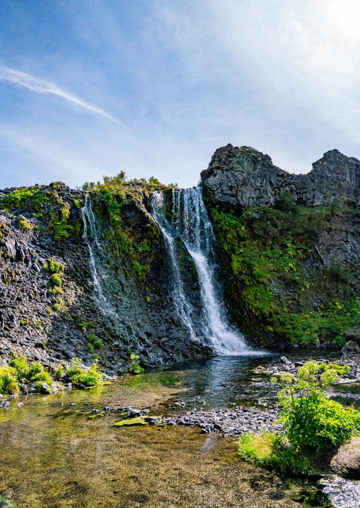 Een waterval stort zich over een rotsachtige, met mos bedekte klif in een heldere, ondiepe poel in IJsland, omringd door groen dat groei symboliseert onder een helderblauwe hemel.