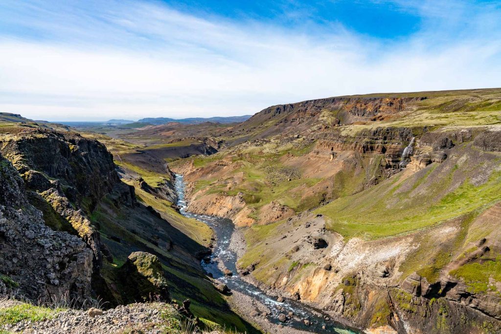 Een weids uitzicht op een rotsachtige canyon in IJsland waar een kronkelende rivier doorheen stroomt, omringd door groene groeiplekken en ruige heuvels onder een blauwe lucht met lichte wolken.