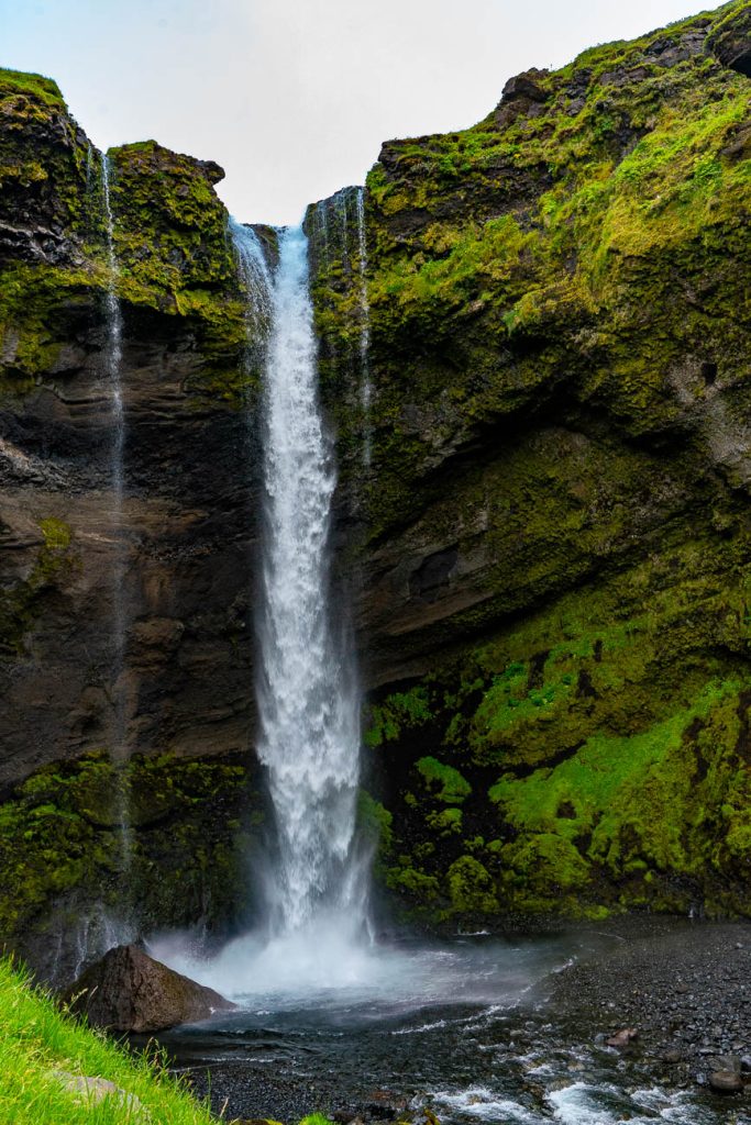 Een hoge waterval stort langs een met mos bedekte klif naar beneden in een poel, omringd door een weelderige groene vegetatie en rotsen. De meestal bewolkte hemel verraadt de wilde schoonheid van IJsland.