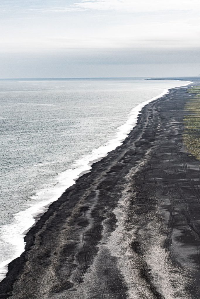 Een lang stuk zwart zandstrand in IJsland ontmoet de grijze oceaan onder een bewolkte hemel, met vage bandensporen zichtbaar op de donkere oever en schaars groen dat aan de rechterkant groeit.