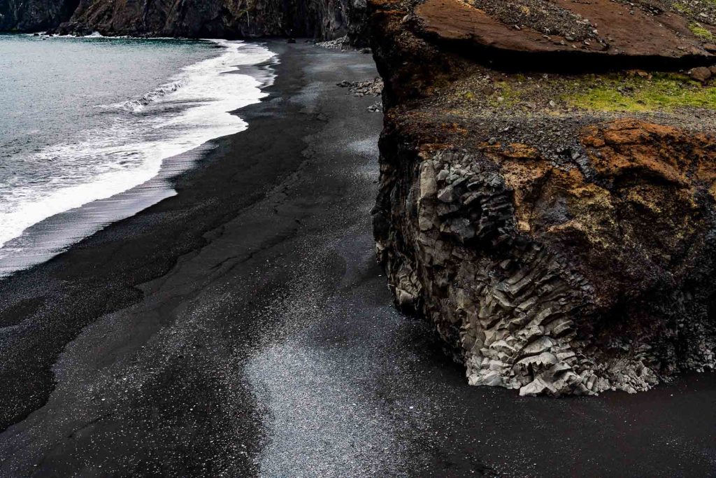 Een ruige klif rijst op boven een zwart zandstrand in IJsland, met donker vulkaanzand dat zich uitstrekt langs de kust en golven die zachtjes aanspoelen, en de rauwe schoonheid en gestage groei van de natuur laten zien.