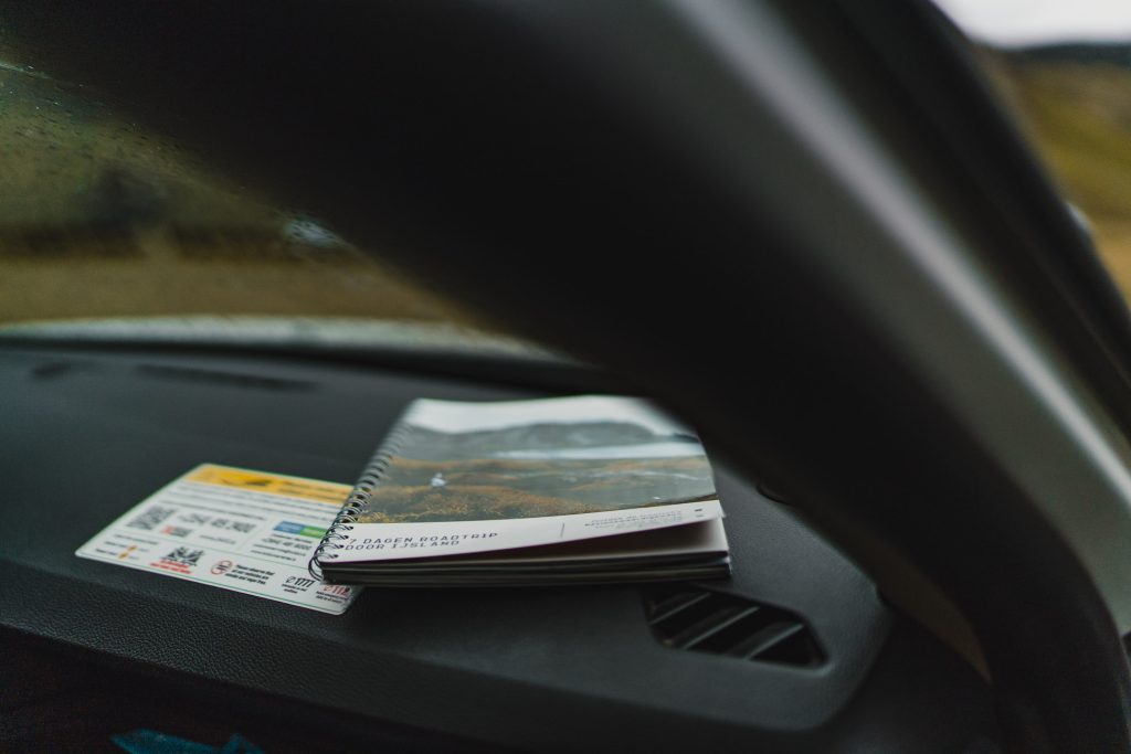Een spiraalgebonden notitieboek met een prachtige foto van een IJsland-landschap op de omslag ligt op het dashboard van een auto, gedeeltelijk in de schaduw van de omlijsting van de voorruit. Naast het notitieboek zijn waarschuwingslabels zichtbaar die groei en avontuur symboliseren voorbij het wazige landschap buiten.
