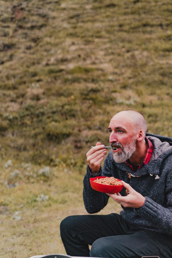 Een man met een grijze baard zit buiten op het gras in IJsland, houdt een rode kom met eten vast en eet met een vork. Hij draagt een grijze trui en een rood geruit overhemd en lijkt te genieten van zijn maaltijd te midden van de natuurlijke groei om hem heen.