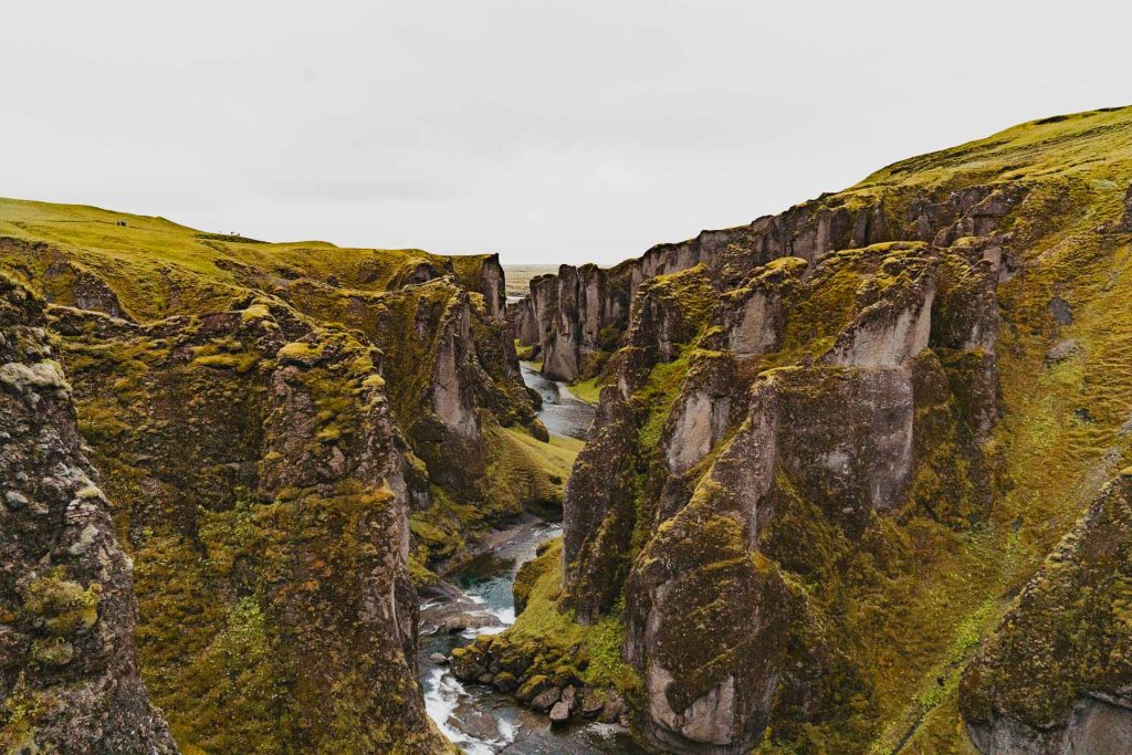Een diepe, smalle canyon met steile, met mos bedekte kliffen en een kronkelende rivier die door het midden stroomt, onder een bewolkte IJsland-hemel - een opvallend landschap waar de groei van de natuur volop te zien is.