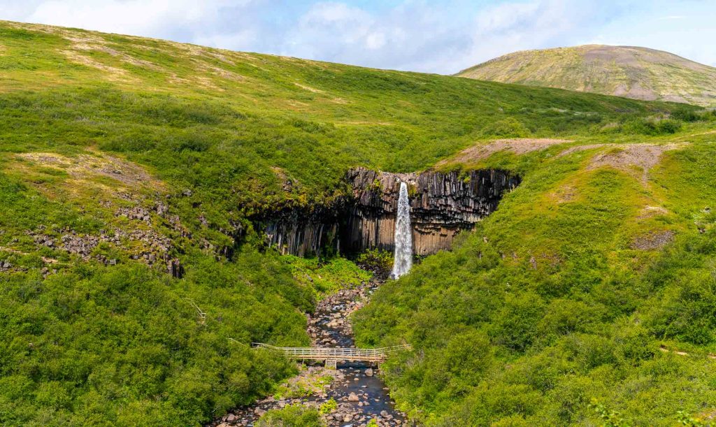 Een hoge, smalle waterval cascadeert over opvallende zuilvormige basaltrotsen in IJsland in een beek, omringd door weelderige groene heuvels die groei belichamen, met een houten loopbrug over het water eronder.