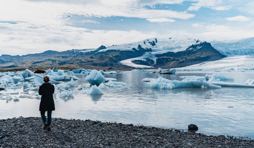 Een persoon in een donkere jas staat op een rotsachtige kust in IJsland en kijkt uit op drijvende ijsbergen in een gletsjerlagune, omringd door bergen met besneeuwde toppen en een bewolkte hemel - een scène die de rauwe schoonheid en groei van de natuur weerspiegelt.