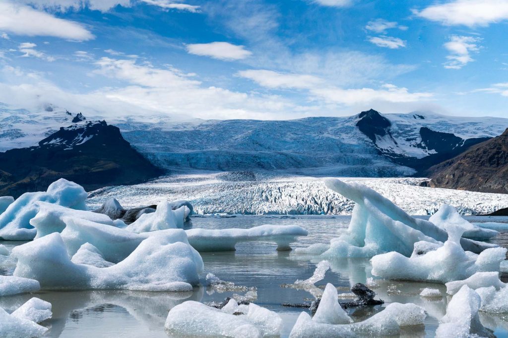 Brokken ijs drijven op een rustig gletsjermeer in IJsland, met een gletsjer en besneeuwde bergen op de achtergrond onder een gedeeltelijk bewolkte blauwe hemel.