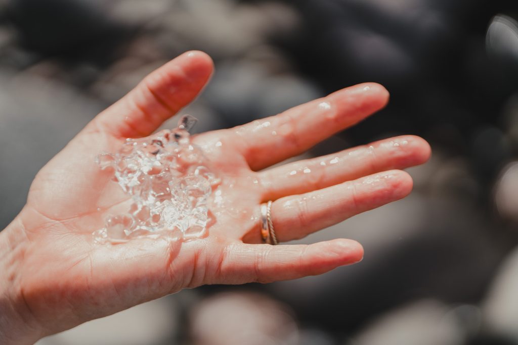 Een close-up van een hand die kleine, heldere stukjes ijs vasthoudt, wat doet denken aan de bevroren schoonheid van IJsland. De natte hand, versierd met een gouden ring aan de ringvinger, steekt af tegen een vage achtergrond met donkere vormen.