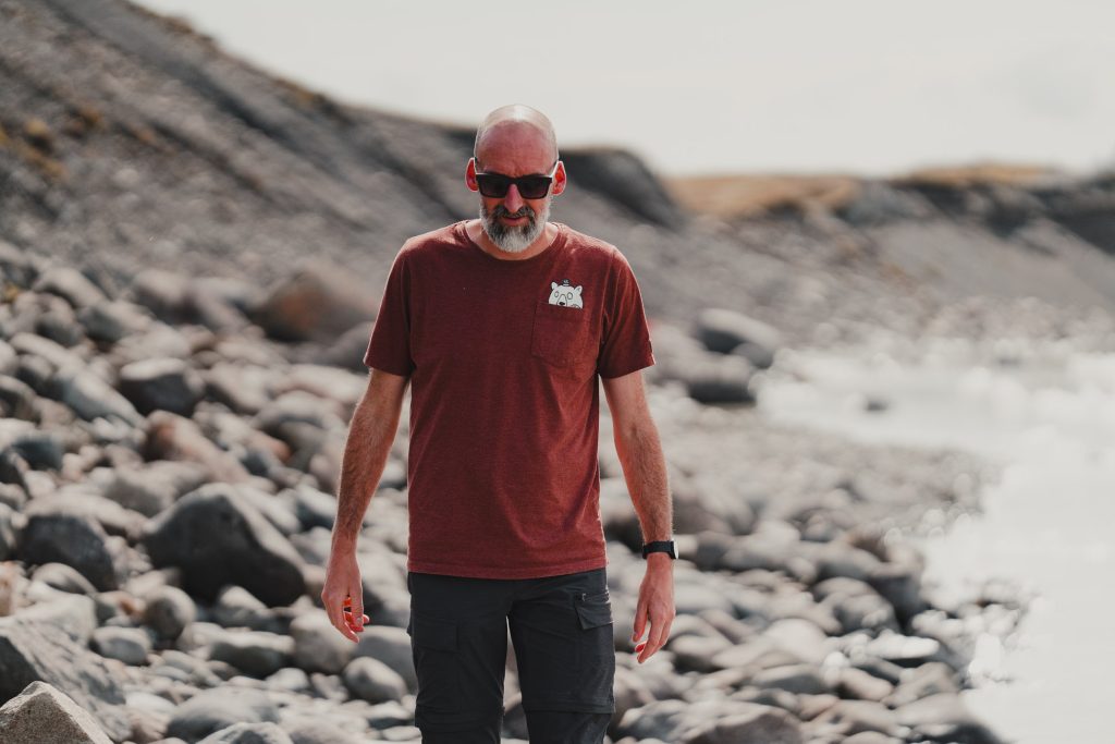 Een kale man met een baard, met een rode zonnebril, een kastanjebruin T-shirt en een donkere korte broek, loopt op een rotsstrand dat eruitziet als IJsland, met vage rotsen en water op de achtergrond waar zijn gevoel voor avontuur groeit.