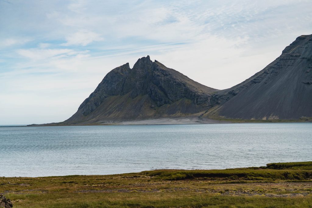 Een ruige berg rijst op naast kalm blauw water onder een lichtbewolkte hemel in IJsland, met groen grasland op de voorgrond dat nieuwe groei suggereert.