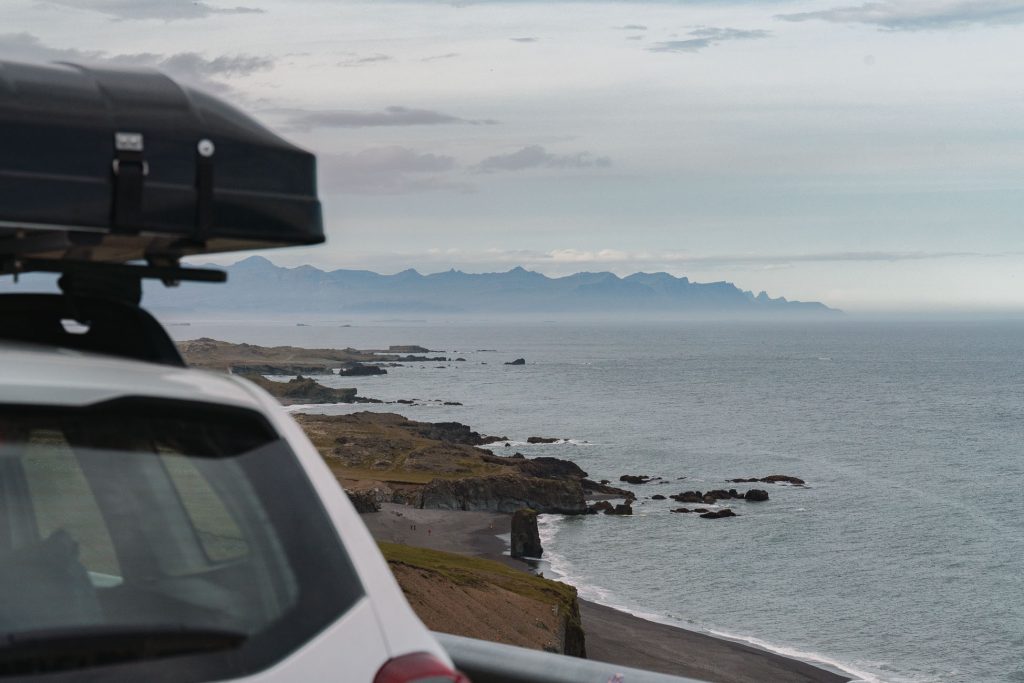 Een witte auto met dakkoffer staat geparkeerd bij een kustklif in IJsland, met uitzicht op een rotsachtige kustlijn en kalme zee met bergen in de verte onder een bewolkte hemel-een perfecte scène voor groei en avontuur.