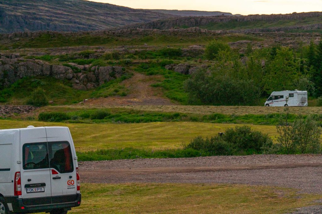 Twee witte campers staan geparkeerd op een grasveld in IJsland, met rotsachtige heuvels en schaars groen op de achtergrond, onder een bewolkte hemel.
