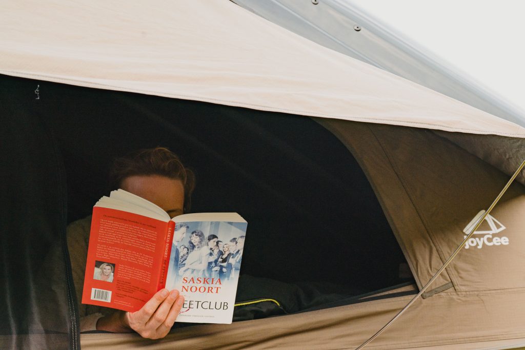 Een persoon zit in een bruine JoyCee tent en leest "De Eetclub" van Saskia Noort. Alleen de ogen en het voorhoofd van de lezer zijn zichtbaar boven het boek, terwijl buiten het landschap de ruige schoonheid van IJsland oproept waar mysteries groeien.