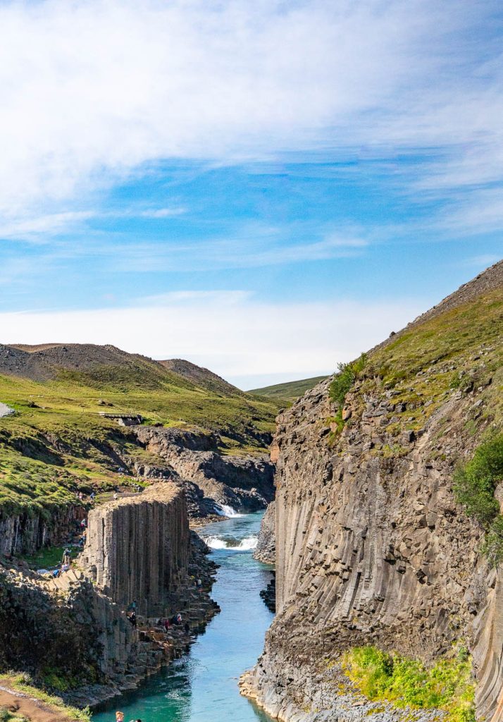 Een helderblauwe rivier stroomt tussen steile, rotsachtige kliffen met duidelijke basaltzuilen in IJsland, omgeven door groene heuvels onder een gedeeltelijk bewolkte hemel. Er zijn mensen te zien die langs de waterkant lopen, ondergedompeld in de serene schoonheid en groei van het landschap.