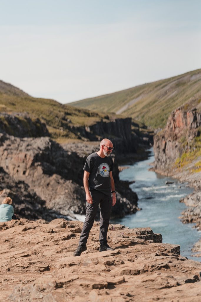 Een man met een baard en een zonnebril staat op een rotsachtig terrein bij een rivier die een groene IJsland-vallei doorsnijdt onder een heldere hemel, die groei symboliseert. Een andere persoon zit op de rotsen op de achtergrond.