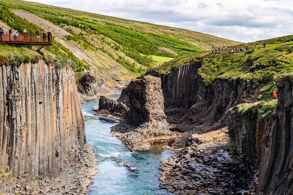 Een rivier stroomt door een canyon met hoge basaltzuilen en groene heuvels in IJsland. Mensen staan op een uitkijkplatform en langs de rand van de klif en bewonderen hoe dit prachtige landschap onder een gedeeltelijk bewolkte hemel groeit.