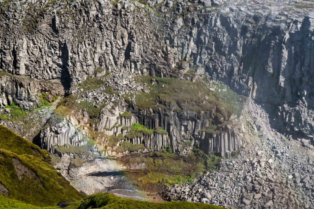 Steile rotsachtige kliffen met zuilen van basaltformaties in IJsland, vlekken groen mos die groei symboliseren en verspreide rotsen aan de voet; linksonder verschijnt een vage regenboog.