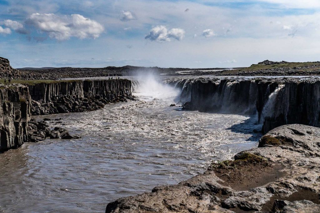 Een brede, krachtige rivier stroomt door rotsachtige kliffen in IJsland en creëert een mistige waterval onder een blauwe hemel met verspreide wolken. Het ruige, meestal kale landschap verraadt de meedogenloze groei en ongetemde schoonheid van de natuur.