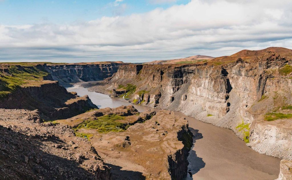 Een brede rivier kronkelt door een diepe, rotsachtige kloof met steile kliffen en schaarse vegetatie onder een gedeeltelijk bewolkte hemel, die doet denken aan de dramatische landschappen waar de natuurlijke schoonheid van IJsland ooit groeide.