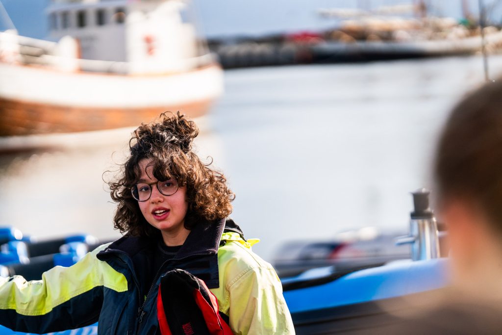 Een jong persoon met krullend haar en een bril, die een geel en zwart jack draagt, staat bij het water met boten op de achtergrond op een zonnige dag in IJsland, een moment van groei en avontuur vastleggend.