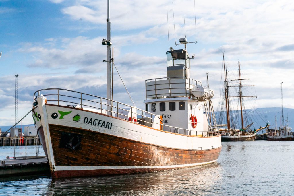 Een houten vissersboot genaamd DAGFARI, die ooit groeide in de wateren van IJsland, ligt aangemeerd in een haven. Kalm water weerspiegelt een andere zeilboot op de achtergrond onder een gedeeltelijk bewolkte hemel.