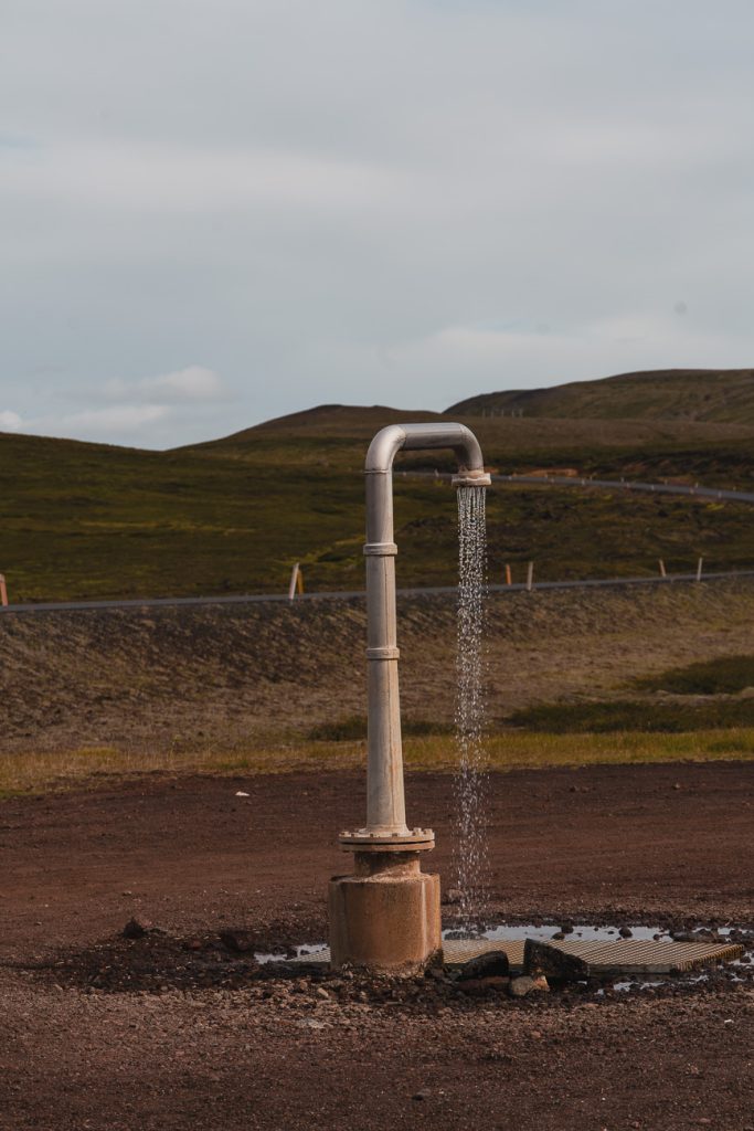 Een metalen pijp staat rechtop buiten in IJsland en laat een gestage stroom water los op de grond met heuvels en graslandschap op de achtergrond onder een bewolkte hemel, groeiend over het terrein.
