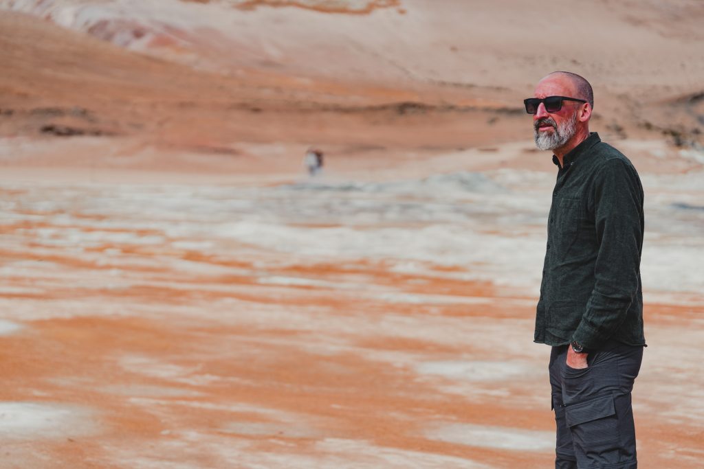 Een man met een zonnebril en donkere kleding staat met zijn handen in zijn zakken op een dor, oranje en wit IJsland-landschap, dat groei symboliseert tegen de grimmige heuvels op de achtergrond.
