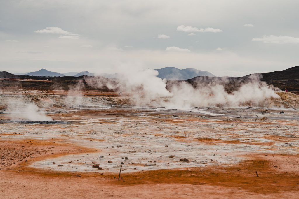 Stoom stijgt op uit geothermische warmwaterbronnen in IJsland, waar een kaal, rotsachtig landschap en bergen in de verte elkaar ontmoeten onder een bewolkte hemel. De grond vertoont oranje en grijze tinten, met plekken van minerale afzettingen die de langzame groei van dit unieke terrein markeren.